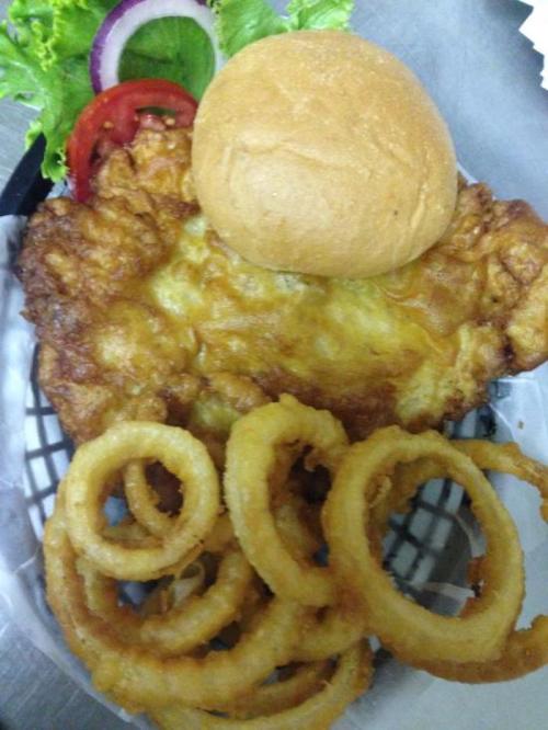A plate featuring a fried chicken fillet, a bun, onion rings, lettuce, tomato, and a slice of onion.