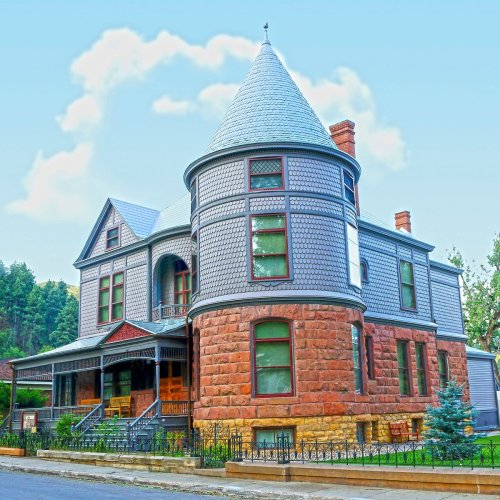 A large, historic house with a turret, featuring a mix of stone and wood siding, surrounded by greenery and a clear sky.