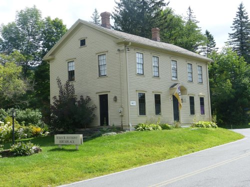 A yellow two-story house with a porch, surrounded by greenery, located on a quiet road.
