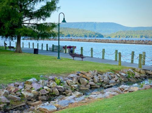 A serene waterfront scene with a wooden dock, green grass, and mountains in the background under a clear blue sky.