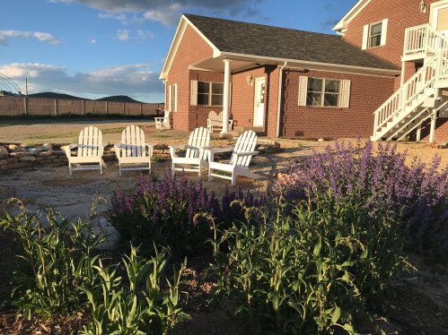 A cozy outdoor space with white Adirondack chairs, purple flowers, and a brick house under a blue sky.