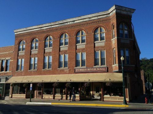 Historic brick building with large windows, featuring "Turkey River Mall" sign and hanging flower baskets. Clear blue sky.