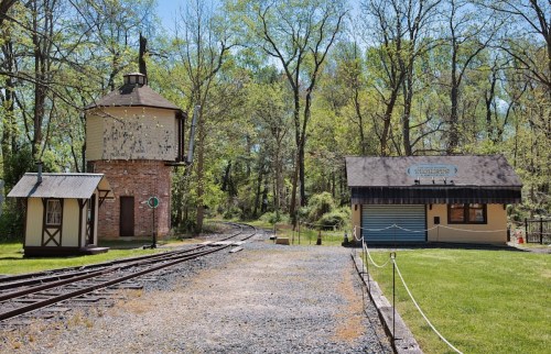 A scenic view of a railway station with a water tower and a building, surrounded by lush greenery and a clear blue sky.