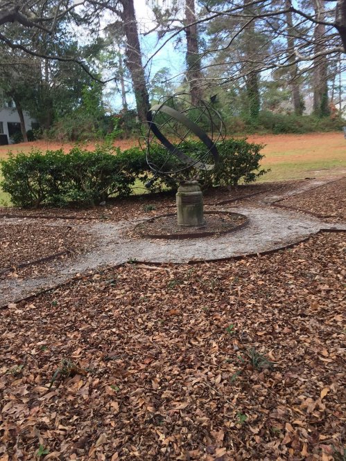 A garden scene featuring a stone pedestal with a metal armillary sphere, surrounded by a circular path and fallen leaves.