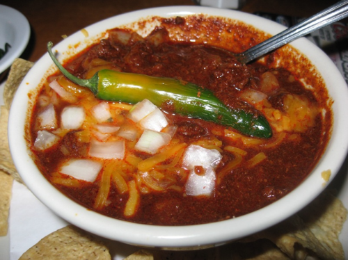 A bowl of chili topped with diced onions, cheese, and a green pepper, served with tortilla chips on the side.