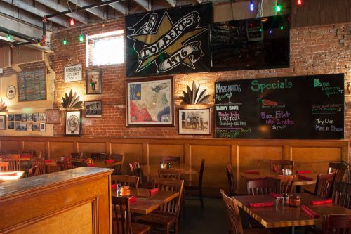Interior of a cozy restaurant with wooden tables, colorful lights, and a chalkboard displaying specials and happy hour details.