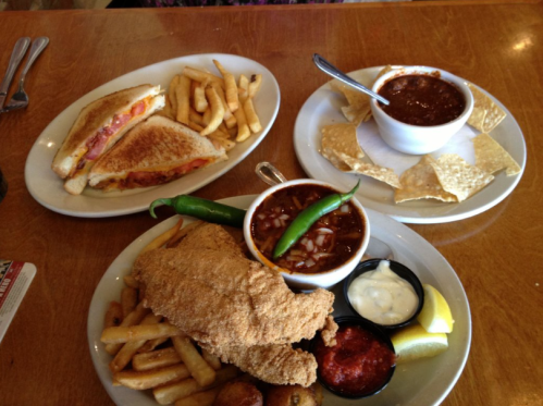 A table with three plates: a sandwich with fries, a bowl of chili with chips, and a fried fish dish with sides.