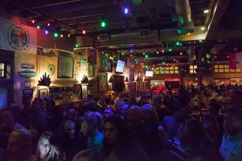 A crowded bar scene with colorful lights, people socializing, and a lively atmosphere.