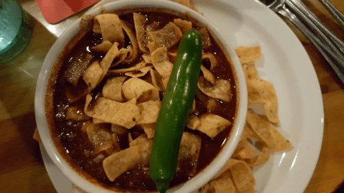A bowl of chili topped with tortilla strips and a green pepper, surrounded by crispy tortilla chips on a plate.
