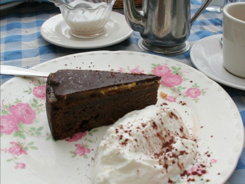 A slice of chocolate cake with a layer of cream, served with whipped cream on a floral plate, alongside a cup and cream jug.
