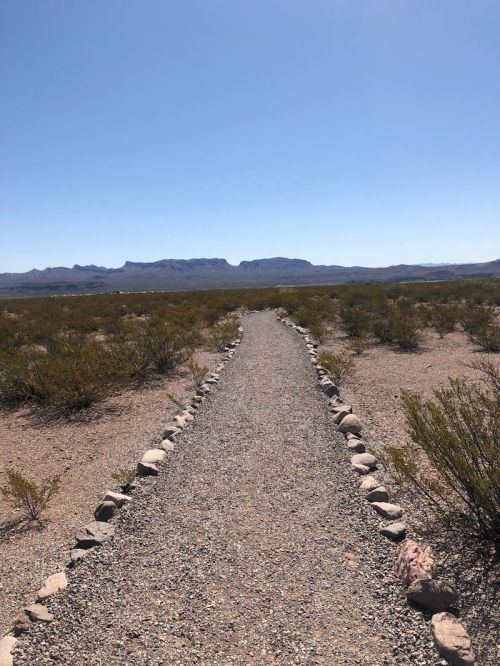 A gravel path lined with stones leads through a desert landscape towards distant mountains under a clear blue sky.