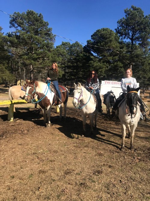 Two women on horseback in a wooded area, smiling and enjoying a sunny day outdoors.