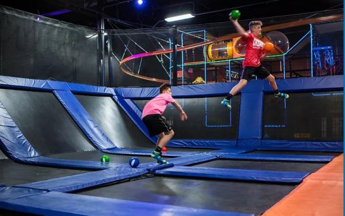 Two boys playing on a trampoline, one jumping in mid-air while the other prepares to throw a ball.
