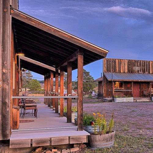 A rustic wooden building with a porch, surrounded by greenery and a blue sky at dusk.