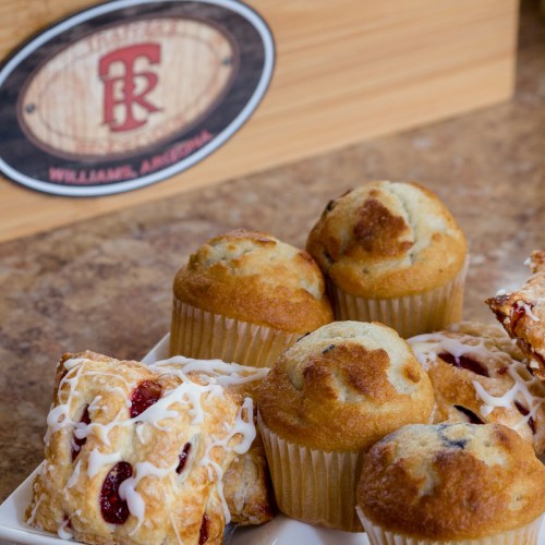 A variety of baked goods, including muffins and pastries, displayed on a wooden surface with a logo in the background.