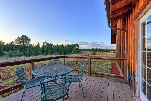 A wooden deck with a round table and chairs overlooks a scenic landscape of trees and open fields under a clear sky.