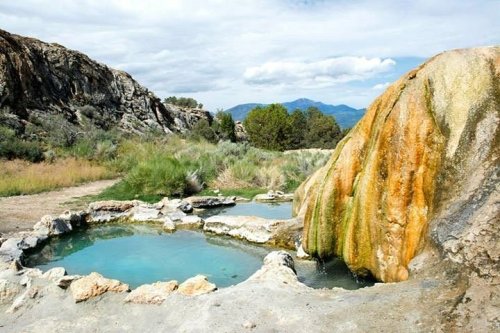 Natural hot springs surrounded by rocky terrain and lush greenery under a cloudy sky.