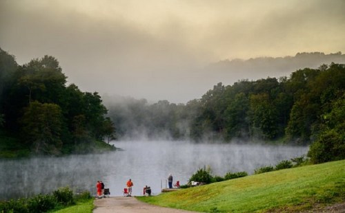 A serene lake scene with mist rising, surrounded by trees and people fishing along the shore in the early morning light.