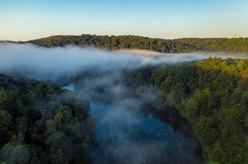 A serene landscape featuring a river surrounded by lush green trees and morning fog under a clear blue sky.