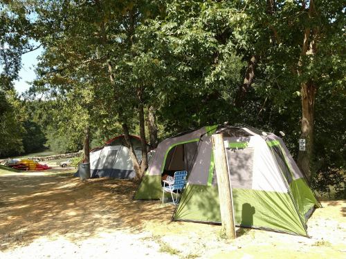 Two tents set up in a wooded area near a river, with a chair and kayaks visible in the background.
