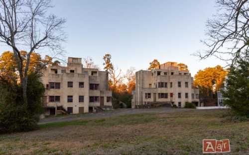 Two abandoned, multi-story buildings with a modernist design, surrounded by trees and a grassy area at sunset.