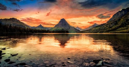 A serene lake reflects a vibrant sunset, surrounded by mountains and lush forests under a colorful sky.