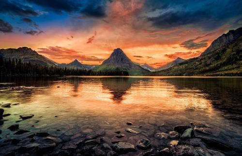 A serene lake reflects a vibrant sunset, surrounded by mountains and lush forests under a colorful sky.