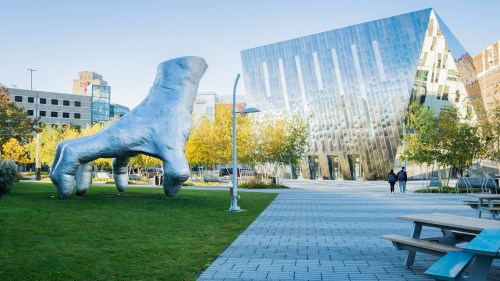 A large, abstract hand sculpture in a park, with a modern glass building and trees in the background.