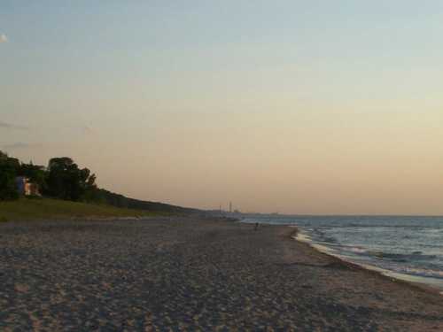 A serene beach at sunset, with soft sand, gentle waves, and a distant shoreline under a colorful sky.