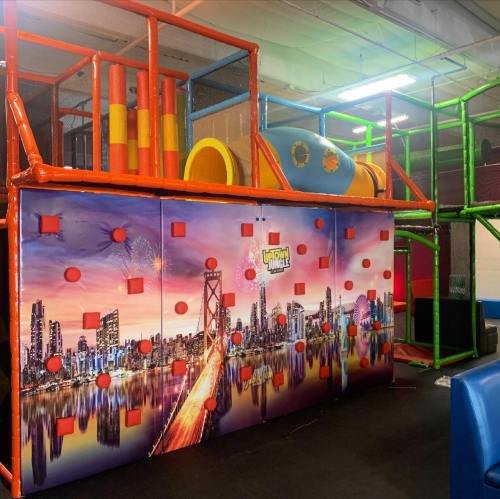 Colorful indoor play structure with climbing walls and tunnels, featuring a city skyline mural in the background.