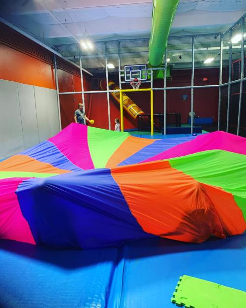 A colorful parachute covers a play area with a basketball hoop and climbing structure in the background.