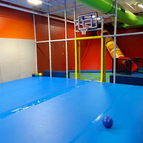 A colorful indoor play area featuring a basketball hoop, slide, and soft play equipment on a blue floor.
