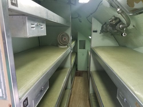 Interior of a narrow room with metal shelves and green bunk beds, featuring a small fan and a door at the end.