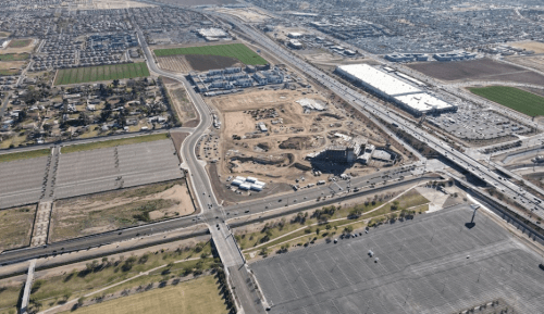 Aerial view of a construction site surrounded by fields, roads, and a large commercial building in the background.