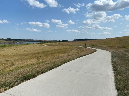 A winding concrete path through a grassy field under a blue sky with fluffy clouds, near a body of water.