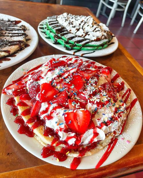 A close-up of a stack of pancakes topped with whipped cream, strawberries, and colorful sprinkles, with two other dessert plates in the background.