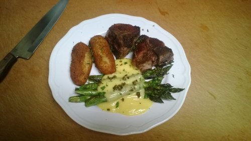 A plate with grilled steak, asparagus, croquettes, and a creamy sauce, arranged on a wooden surface.