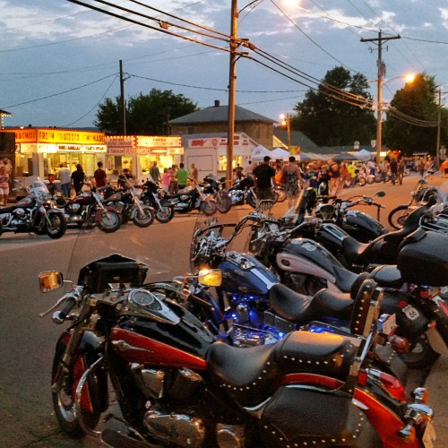 A street scene at dusk with parked motorcycles in the foreground and a lively festival atmosphere in the background.