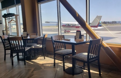 A cozy restaurant with tables by a large window overlooking a small airplane on the runway.