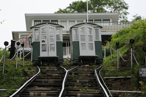 Two vintage cable cars on tracks leading up a hill, with a house and greenery in the background.