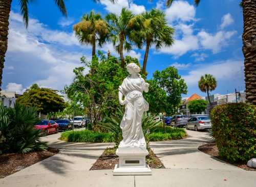 A white statue of a woman stands in a lush garden surrounded by palm trees and parked cars under a blue sky.