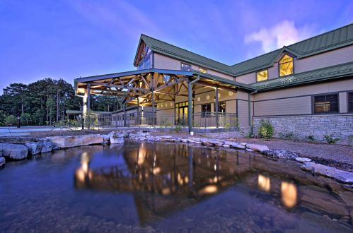 A modern building with a green roof, reflecting in a pond at dusk, surrounded by trees and landscaped grounds.