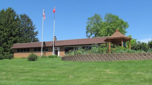 A single-story brick house with two flags in front, surrounded by green grass and a gazebo nearby.