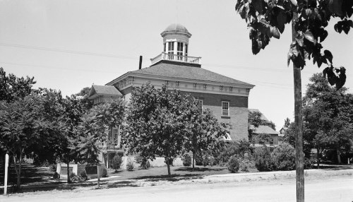 Historic building with a dome, surrounded by trees and greenery, set against a clear sky. Black and white photo.