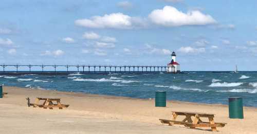 A sandy beach with picnic tables, waves, and a lighthouse on a pier under a partly cloudy sky.