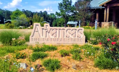 Sign reading "Arkansas The Natural State" surrounded by flowers and greenery at a park entrance.