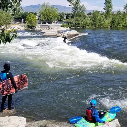 Two people in wetsuits near a river, one on a surfboard and another in a kayak, with trees and mountains in the background.