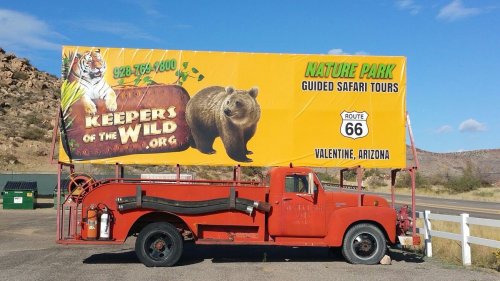 A vintage red truck parked beside a large billboard advertising guided safari tours at Keepers of the Wild in Arizona.
