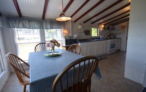 Cozy kitchen with a wooden table, checkered tablecloth, and bright windows, featuring a simple cooking area.