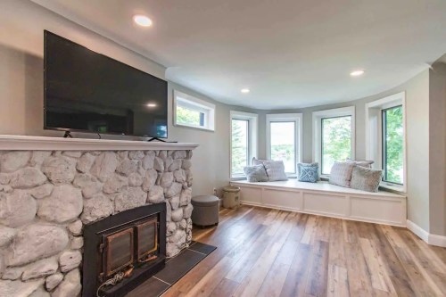 Cozy living room with a stone fireplace, large TV, and bay windows with cushions, featuring wooden flooring.